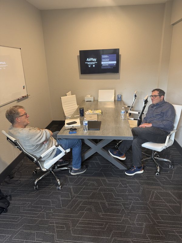 Two people in a meeting in our enclosed conference room, a wall screen showing AirPlay and a whiteboard behind them