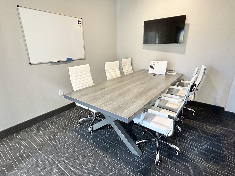 A private conference room with a grey wood table, white chairs, a whiteboard and a wall-mounted screen