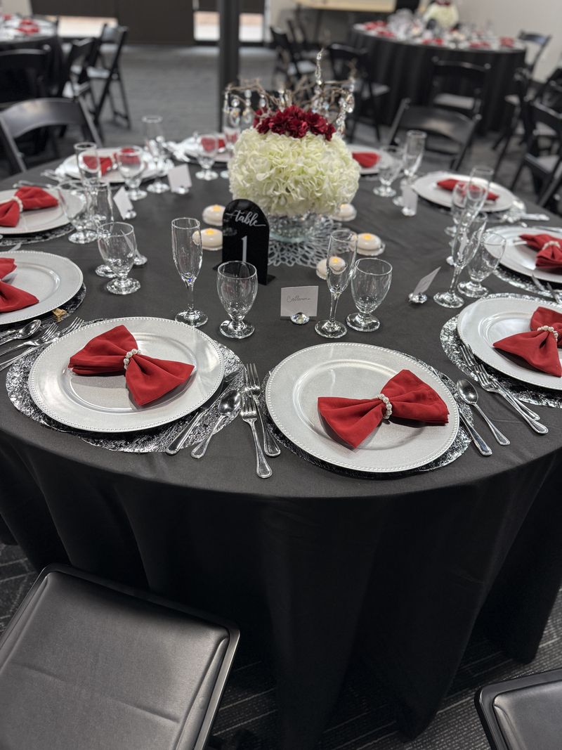 A round table set with silver chargers, red napkins tied in bows, glassware and a white flower centerpiece