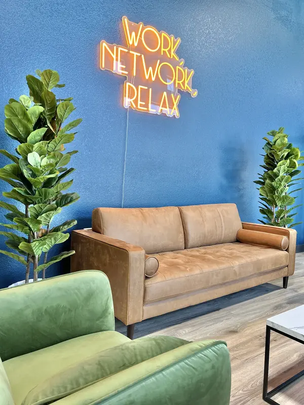 A tan leather sofa against the lounge's navy wall under the Work, Network, Relax neon, with a green velvet chair and two fiddle-leaf figs