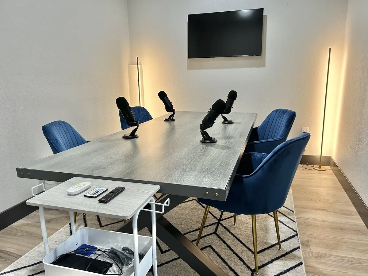 A podcast room set up with four microphones on a table, blue velvet chairs, a wall screen and floor lamps