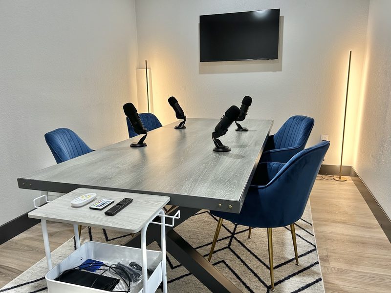 A podcast room set up with four microphones on a table, blue velvet chairs, a wall screen and floor lamps